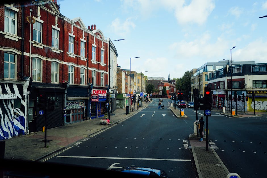 Photograph of a city street scene during daytime, showing a mix of red brick and modern buildings on either side of the road. The street features a vehicle loading and unloading area with a man with van dressed in casual clothing, carrying a large wrapped piece of furniture secured with plastic wrap and moving straps, heading towards a white van parked nearby. Several cardboard boxes and smaller packages are placed on the pavement, indicating a furniture or household item move as part of home relocation services by Man with Van Pentonville. The street has traffic lights, pedestrian crossings, and a few pedestrians waiting at the curb, with some parked cars visible in the background. The lighting is bright, and the sky is partly cloudy, providing clear visibility of the moving process within the urban environment, reflecting professional removal and furniture transport activities consistent with house removals and packing and moving logistics.