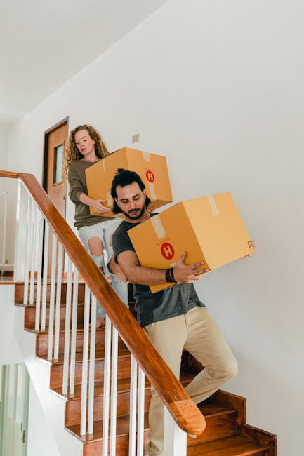 A woman with shoulder-length brown hair, wearing a beige jacket, is carrying a large cardboard box as part of the furniture and home relocation process inside a narrow staircase of a residential property. She is stepping down the staircase, which is lined with dark wood flooring, while holding the box securely. Behind her, a person with short hair, dressed in a blue and black checkered shirt, is also involved in the moving task, possibly assisting with furniture transport. The staircase features white walls and a black metal handrail on the right side, with natural light coming in from a window at the top of the stairs. Visible moving materials include additional cardboard boxes and possibly packing blankets or wraps used to protect furniture during transport. The setting is within an indoor hallway, supporting the logistics of packing and loading items onto a vehicle for home relocation, as handled by Man with Van Pentonville.