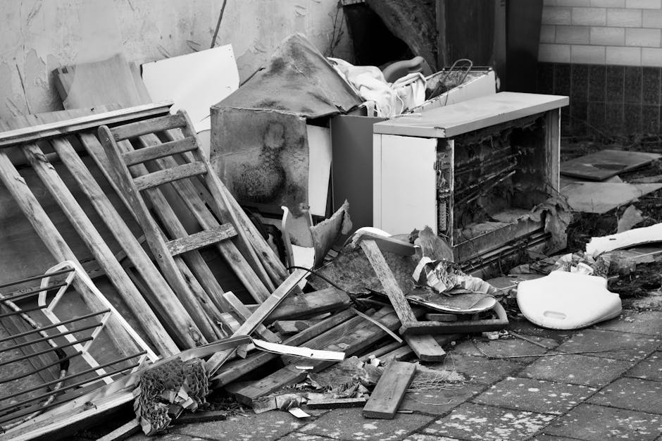 A black-and-white photograph showing an outdoor scene of household rubbish and discarded furniture placed on a pavement outside a building. The items include a disassembled wooden bed frame leaning against a wall, several cardboard boxes with contents spilling out, a broken office chair, a damaged old microwave or appliance, and various pieces of broken wood and plastic debris scattered on the ground. The background features a brick and tiled wall, with some fabric or padding draped over a metal frame or trolley. The scene suggests removals or clearance activity, with clutter likely awaiting disposal or collection by a professional moving or clearance service, such as Man with Van Pentonville, as part of house removal or property clearing processes.