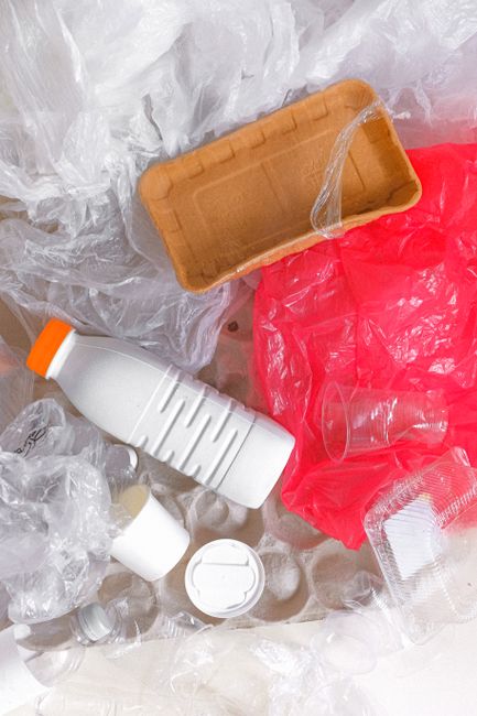 A collection of various discarded waste and packaging materials including crumpled white and red plastic bags, transparent plastic bottles, a cardboard food container, a plastic drinking cup with a straw, and a plastic lid, all scattered on a surface likely during a home relocation packing process. The background consists of white tissue or bubble wrap, indicative of household packing materials. These items are surrounded by loose plastic debris, suggesting the decluttering or trash removal stage of a house move. The setting appears to be indoors, with no visible furniture or people, and the scene highlights the importance of proper waste disposal and recycling in the context of a house removals service. Man with Van Pentonville provides moving and packing solutions suited for handling such items during a home relocation in the Pentonville area, aligning with regulations on bulky-item disposal and move logistics.