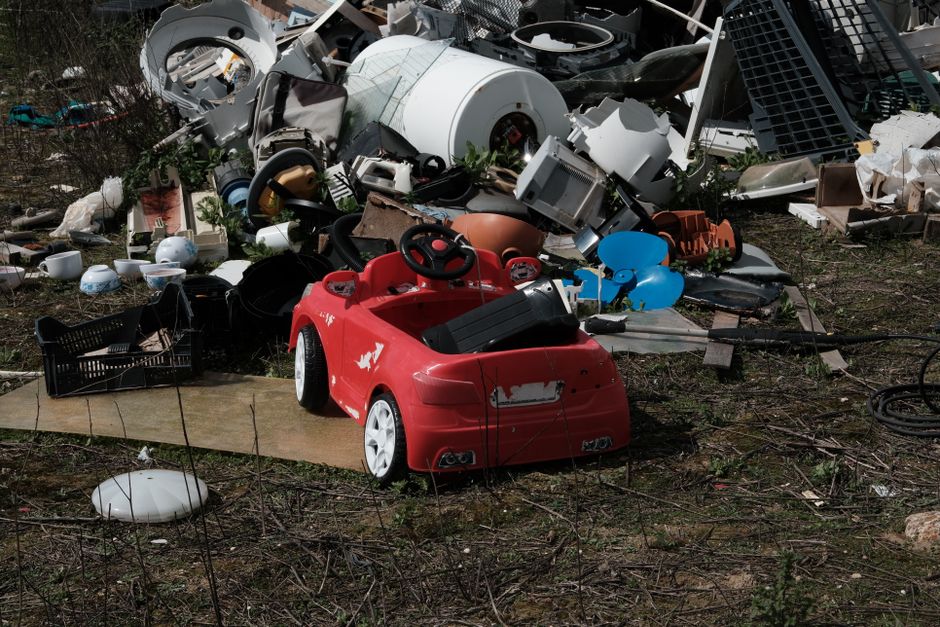 A black-and-white photograph showing an outdoor scene of household rubbish and discarded furniture placed on a pavement outside a building. The items include a disassembled wooden bed frame leaning against a wall, several cardboard boxes with contents spilling out, a broken office chair, a damaged old microwave or appliance, and various pieces of broken wood and plastic debris scattered on the ground. The background features a brick and tiled wall, with some fabric or padding draped over a metal frame or trolley. The scene suggests removals or clearance activity, with clutter likely awaiting disposal or collection by a professional moving or clearance service, such as Man with Van Pentonville, as part of house removal or property clearing processes.