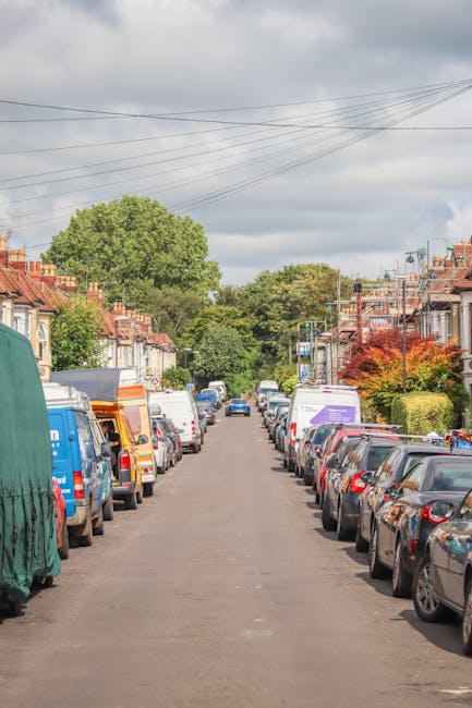 A quiet residential street with parked cars lining both sides of the road, including hatchbacks, sedans, and vans, with some covered by protective tarps. At the far end, a man with a van, part of the 