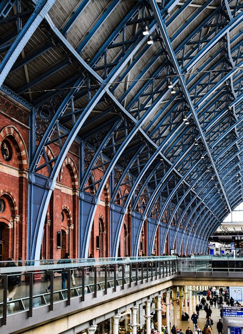 Interior view of a large train station with a high, arched ceiling supported by blue steel beams and trusses. The ceiling features a grid of skylights allowing natural light to illuminate the space. Red brick walls with arched windows line the upper level, where a glass and metal railing runs along the edge of the walkway. On the lower level, numerous travelers, some with luggage and boxes, walk through the busy station. In the foreground, a section of a van or transport vehicle is visible on a paved area, likely used for home relocation or furniture transport, with the parking area and station signage in the background. The image captures the moment of loading or unloading items during a house removal or professional moving process, showcasing the logistical environment of a busy transit hub as part of a furniture transport operation coordinated by Man with Van Pentonville.