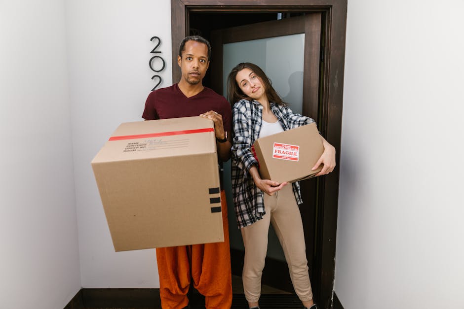 A man and a woman standing inside a residential doorway, each holding cardboard boxes used for home relocation. The man, dressed in a maroon shirt and loose orange pants, holds a large box with black tape and a red-striped shipping label visible on top, indicating packing materials for furniture transport. The woman, wearing a plaid shirt over a white top and beige pants, carries a smaller cardboard box marked with 'FRAGILE' labels. They are positioned near an open door with the house number visible to the left, inside an entryway with white walls and a dark-colored floor. The scene depicts the loading process, as they prepare to move boxes into or out of the property during a professional house removal or packing and moving service. The environment is well-lit, emphasizing the focus on the boxes and the moving activity, consistent with the services of Man with Van Pentonville specialized in urgent flat moves and same-day removals.