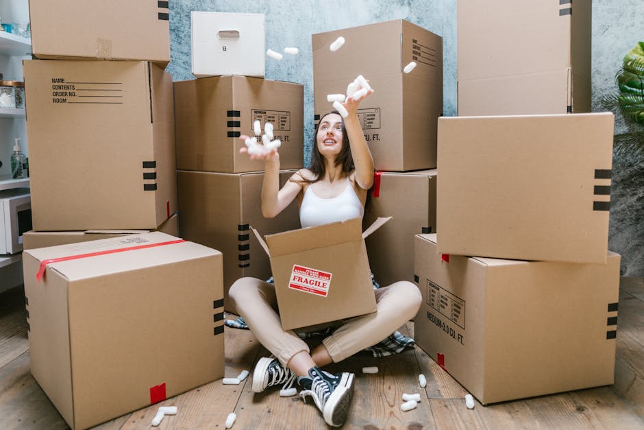 A young woman with long dark hair, wearing a white tank top, beige pants, and black and white sneakers, is sitting on a wooden floor inside a room surrounded by large cardboard moving boxes, some sealed with red tape and others open, revealing packing materials like foam peanuts. She is smiling and looking upwards while tossing foam packing peanuts into the air. The boxes are stacked and arranged around her, with some on her sides and one partially off her lap, showing a 'Fragile' sticker on it. Behind her, there is a wall with blue and gray textured paint, a small plant in the corner, and a glimpse of a white cabinet or shelf to the left. The scene depicts a home relocation or packing process, with natural and indoor lighting emphasizing the casual, preparatory atmosphere associated with furniture transport and house removals. This image relates to services provided by Man with Van Pentonville, specializing in relocations and moving logistics.