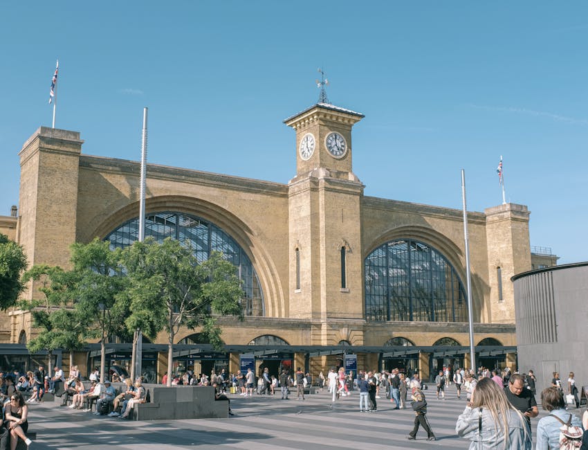 A large historic train station with a beige brick facade, featuring a central clock tower with a white clock face and decorative elements at the top, situated above arched glass windows. The station is busy with a crowd of people walking across the open outdoor area in front of the building, some seated on benches and others standing or walking. In the foreground, there are two flagpoles without flags, and the scene is illuminated by bright daylight under a clear blue sky. Green trees partially obscure the lower part of the station, and the area appears to be a popular travel hub, with considerations relevant to house relocations and furniture transport. The image highlights the environment where professional removals by Man with Van Pentonville may take place, focusing on loading or unloading logistics in a busy urban setting near King's Cross, associated with moving and packing processes.
