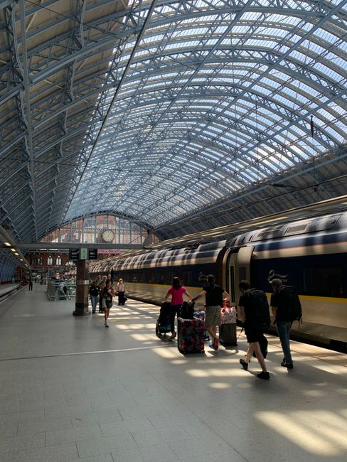 Inside a large railway station with an arched glass roof supported by metal framework, featuring a prominent round clock on a metal lattice wall and a pink neon sign with cursive text. The station flo