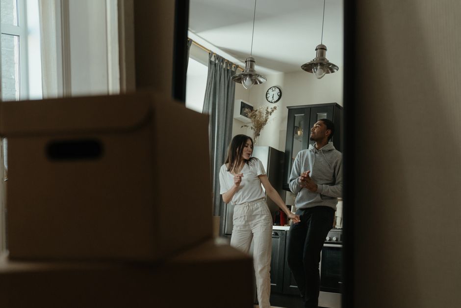 A view through a mirror showing a young woman and a young man inside a kitchen during a house removal process. The woman has long dark hair and is wearing a white T-shirt and beige trousers, while the