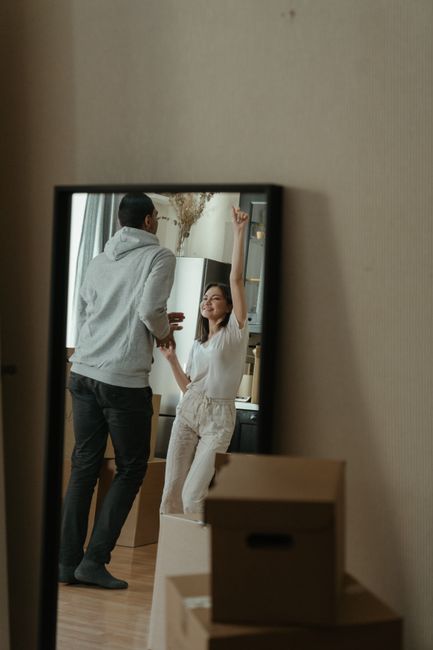 A view through a mirror showing a young woman and a young man inside a kitchen during a house removal process. The woman has long dark hair and is wearing a white T-shirt and beige trousers, while the