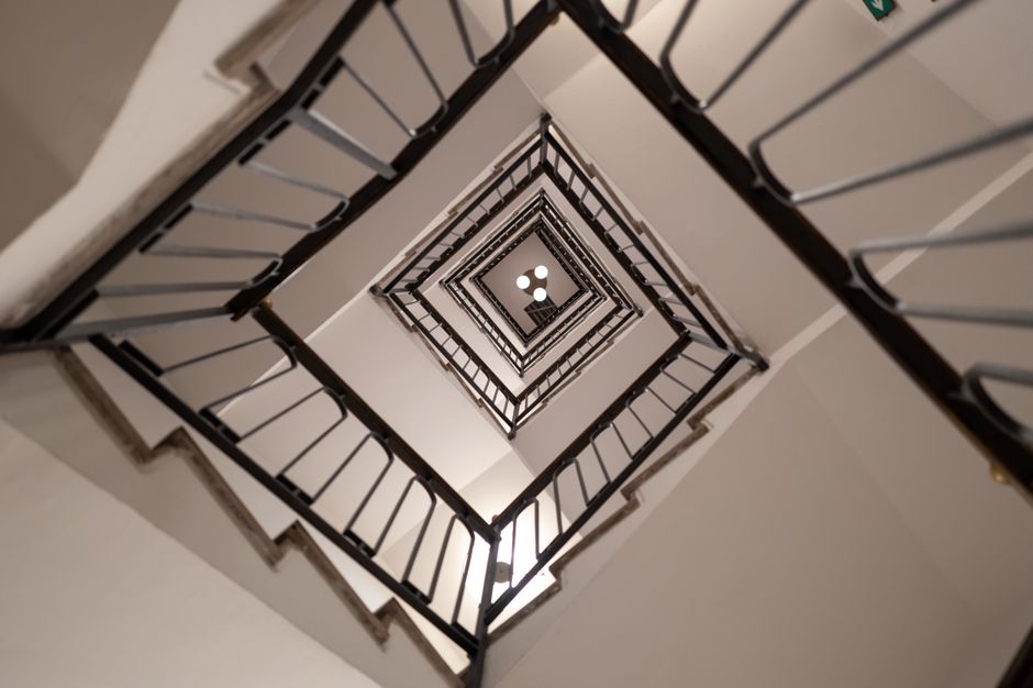 A view looking directly down the central opening of a multi-storey staircase in an interior residential setting, capturing the square-shaped landings on each floor with black metal railings and white walls. The staircase's design includes a series of enclosed, symmetrical levels that spiral downward, with the vertical section visible through the angular framing. The lighting illuminates the stairwell evenly, highlighting the clean lines and the contrast between the black railing and white interior walls. This image likely reflects a home with internal staircases, possibly Victorian or period architecture, which poses challenges for furniture transport during a home relocation. The scene illustrates the complexity of moving large items through narrow or winding staircases, which is relevant when considering professional removals services, such as those offered by Man with Van Pentonville, especially for tight corridors or narrow stairwells in the N1 area.