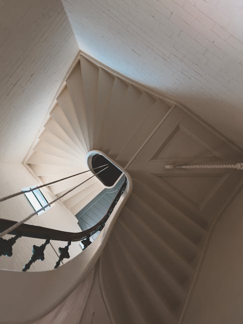 A narrow, curved staircase inside a residential property with white-painted wooden banisters and a dark, ornate handrail. The stairs are carpeted in a light fabric. At the top of the staircase, an open doorway reveals a glimpse of a small window allowing natural light into the space. The staircase is surrounded by white-painted brick walls. This interior scene is part of a home undergoing a professional removal or moving process, such as packing and furniture transport coordinated by Man with Van Pentonville, to facilitate house relocation despite confined and challenging Victorian staircase dimensions.