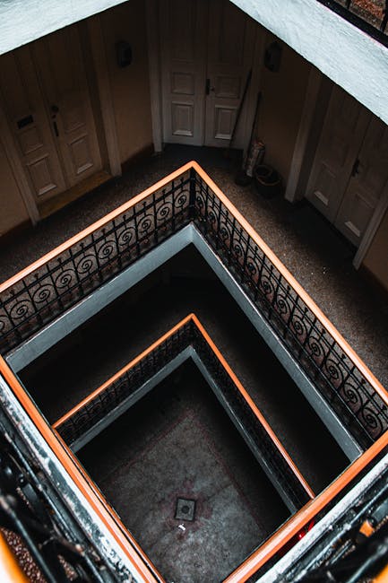 A view looking directly down the central opening of a multi-storey staircase in an interior residential setting, capturing the square-shaped landings on each floor with black metal railings and white walls. The staircase's design includes a series of enclosed, symmetrical levels that spiral downward, with the vertical section visible through the angular framing. The lighting illuminates the stairwell evenly, highlighting the clean lines and the contrast between the black railing and white interior walls. This image likely reflects a home with internal staircases, possibly Victorian or period architecture, which poses challenges for furniture transport during a home relocation. The scene illustrates the complexity of moving large items through narrow or winding staircases, which is relevant when considering professional removals services, such as those offered by Man with Van Pentonville, especially for tight corridors or narrow stairwells in the N1 area.